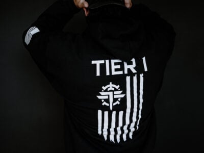 Back view of a person in a black hoodie with 'TIER 1' graphic on the back, hands on a camouflage cap, dark background.