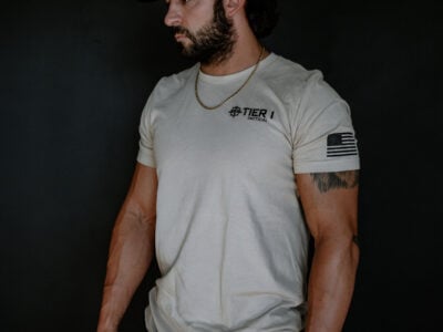 Bearded man in a beige Tier 1 Tactical T-shirt, camo cap, and gold chain, facing left against a dark background.